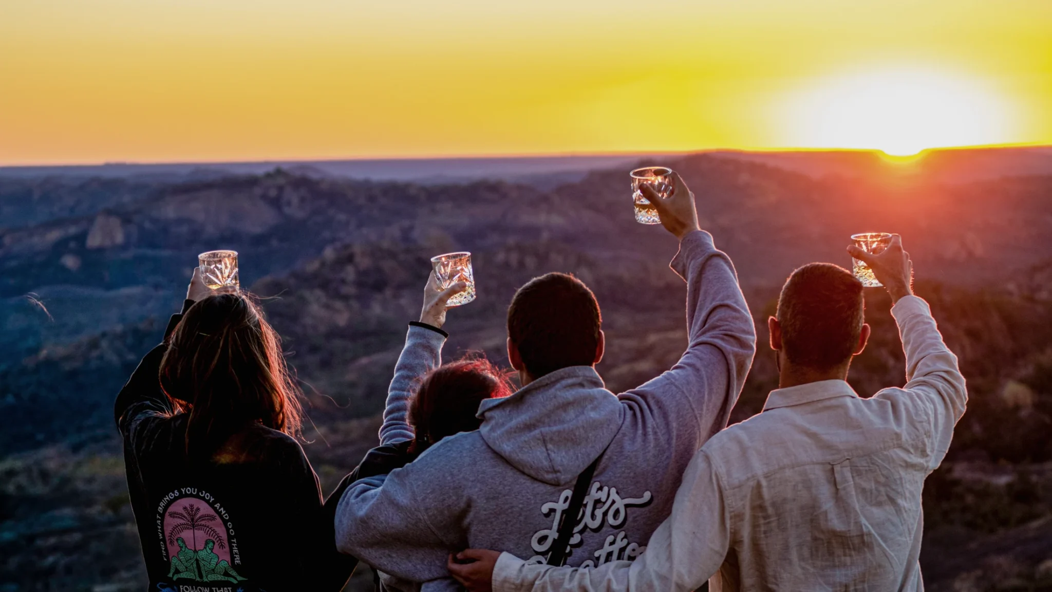 Four friends raising their glasses towards the setting sun on the summit of World’s View